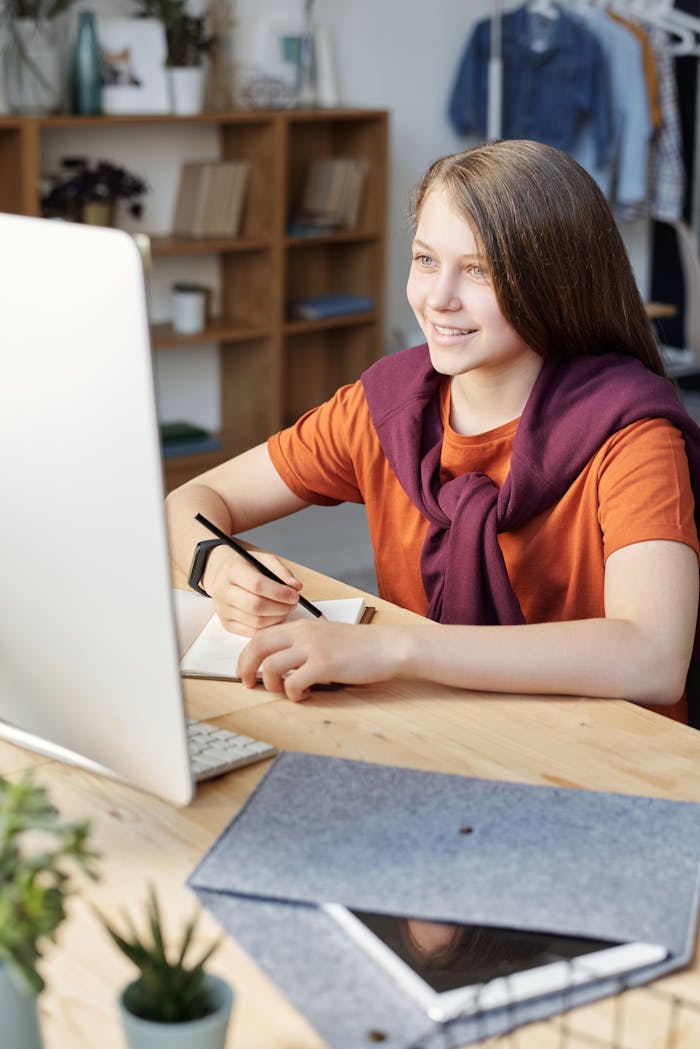 our-story Smiling teen girl studying online at her home desk with a computer and notepad.