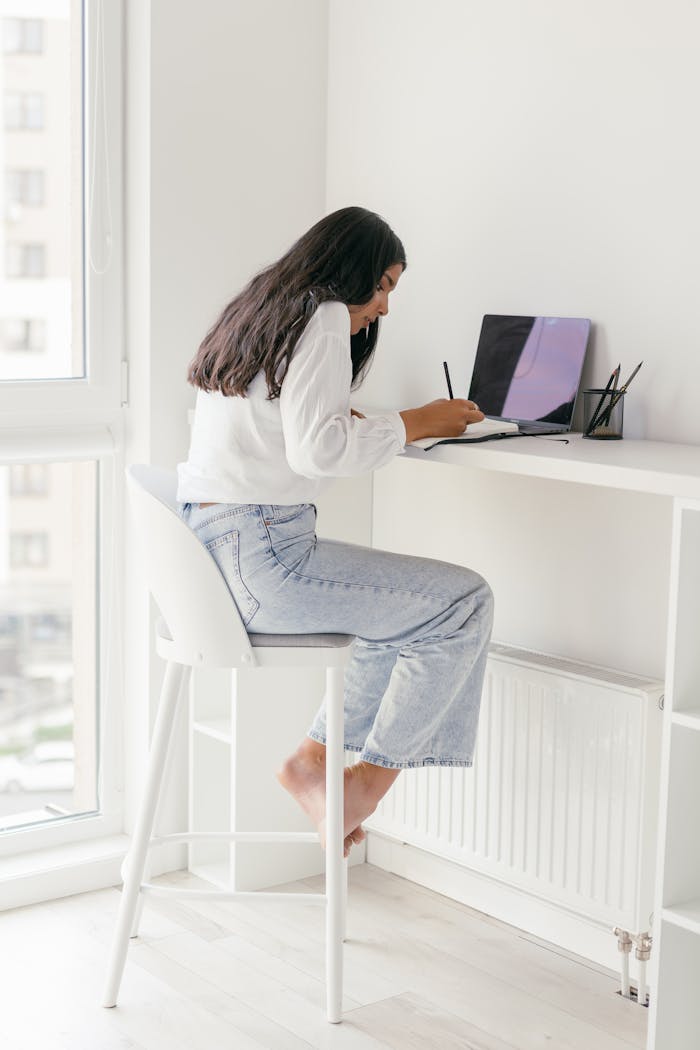 A young woman sits at a desk, focusing on her laptop in a bright home setting, ideal for remote work.